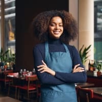 Smiling female restaurant worker wearing an apron, standing confidently with arms crossed in a modern dining area filled with plants and tables. This image highlights the welcoming atmosphere of the restaurant, ideal for illustrating customer service or hospitality topics.
