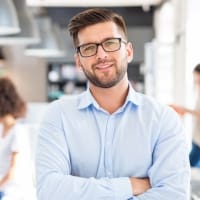 Smiling young professional man in a blue shirt and glasses, standing confidently in a modern office environment, representing a positive workplace culture and professional development.
