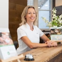 Smiling female receptionist in a healthcare clinic setting, welcoming patients at the front desk with a wooden counter and informational brochures nearby.
