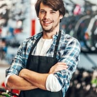Smiling young male shopkeeper in a plaid shirt and apron, standing confidently in a bicycle store, surrounded by bicycles and accessories. Ideal for a page about local bike shops or customer service in retail.