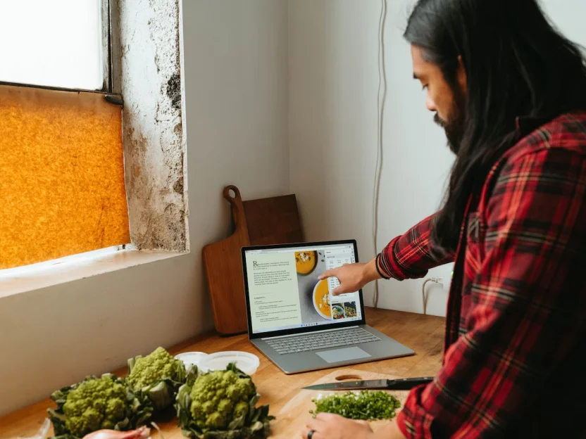 A person in a red flannel shirt preparing a meal in a cozy kitchen, using a laptop to follow a recipe while surrounded by fresh ingredients like greens and grapes.