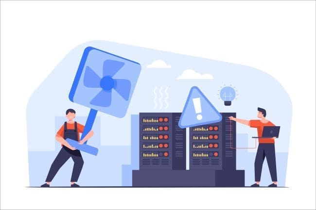 Two technicians troubleshooting a server room, one holding a large fan to cool equipment while the other monitors alerts on a laptop. This image illustrates the importance of maintaining optimal temperatures in data centers to prevent overheating.